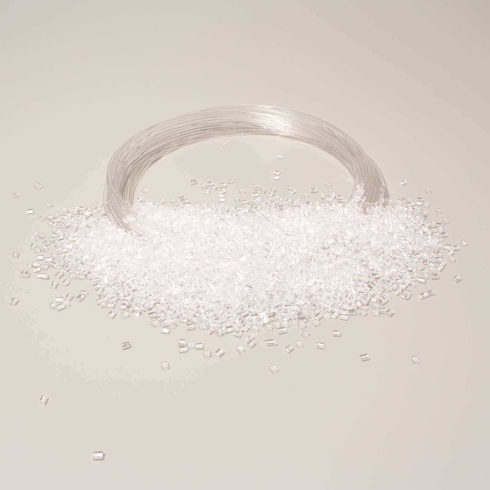 Granuli trasparenti di plastica insieme a un filamento dello stesso materiale su sfondo chiaro.