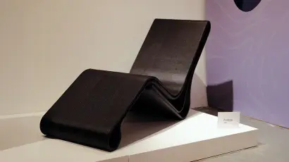 Sedia a sdraio moderna dal design curvo in mostra, realizzata con materiale scuro, su piattaforma bianca.